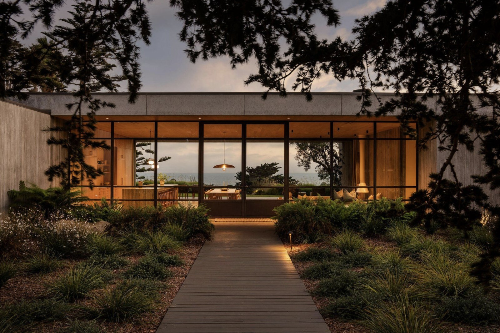 Boardwalk path through native grasses to the glass entry of 3150 Sea Cliff at dusk, with the ocean visible through the house