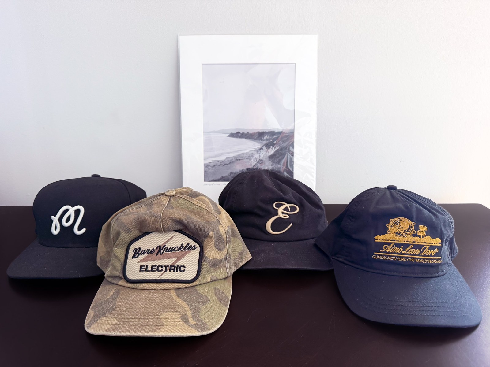 Four hats from Malbon, Bare Knuckles, Eliora, and Aimé Leon Dore, lined up on a dresser